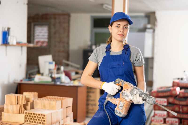 Smiling young female builder in blue overalls and cap standing at construction site