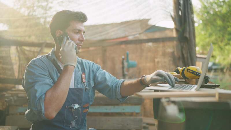 Young male carpenter using smartphone and laptop