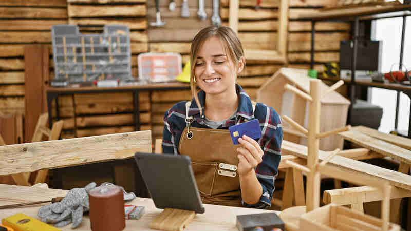 blonde woman carpenter smiling while paying with credit card in carpentry workshop
