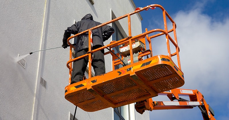 Worker on platform washing building exterior with pressure washer