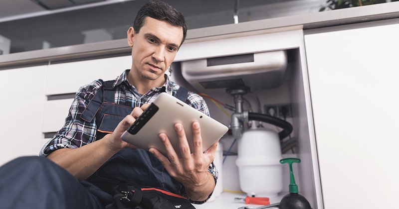 Plumber sits next to kitchen sink on floor and looks at tablet