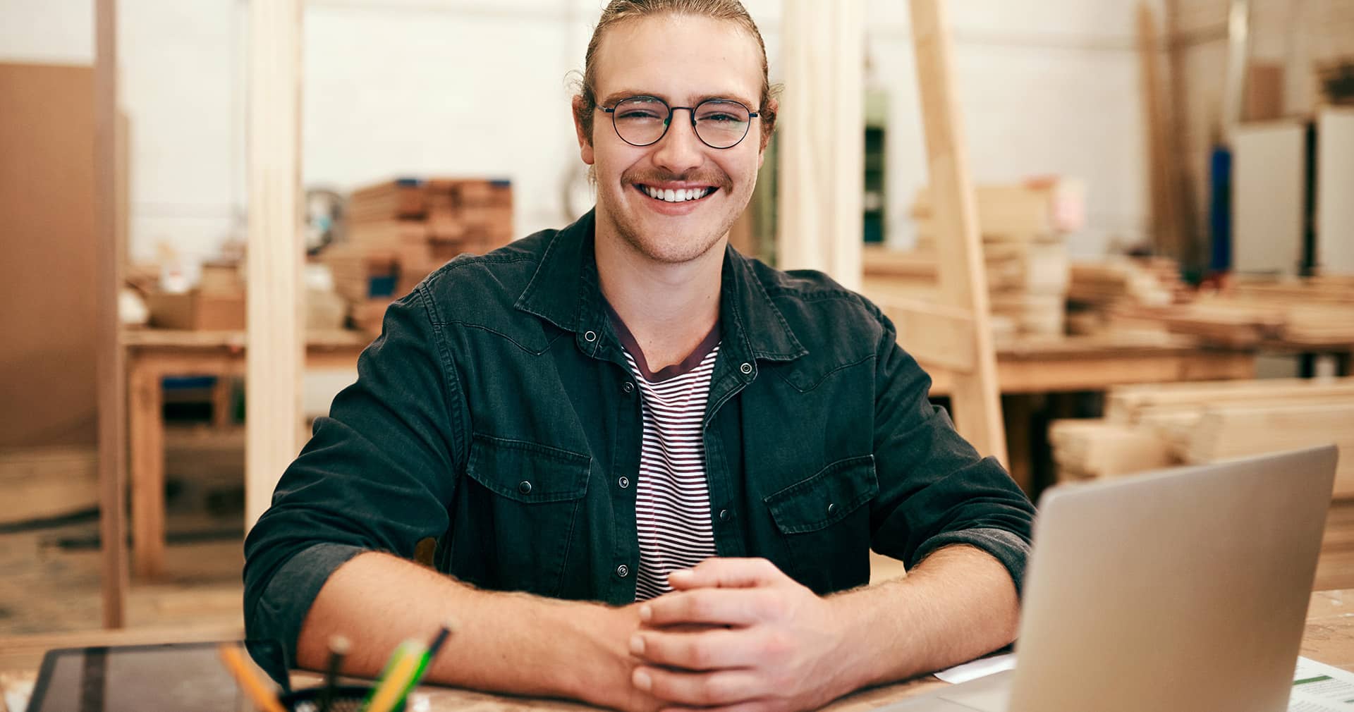 Carpenter in workshop in front of open laptop, smiling at camera