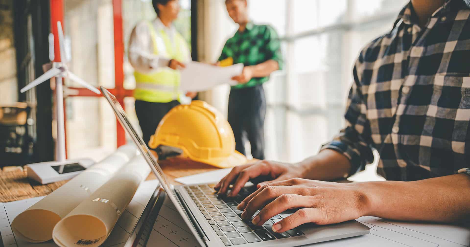 Closeup of contractor's hands typing on open laptop with blue print and yellow helmet on desk