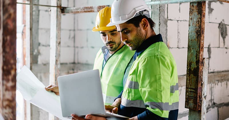 Two construction workers at building site looking at blue print and laptop