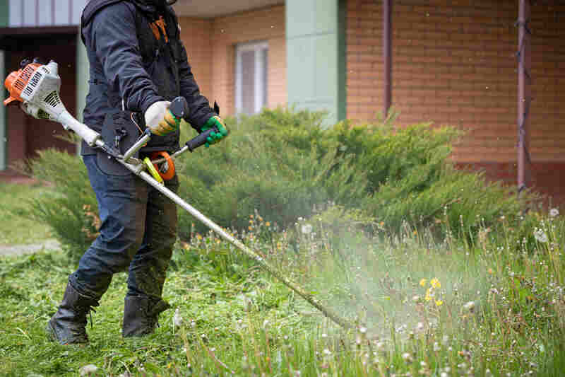 A man in protective clothing, gloves and rubber boots mows the grass with a trimmer