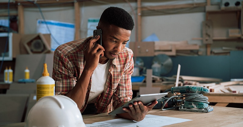 Carpenter in workshop on phone and reviewing estimate on tablet