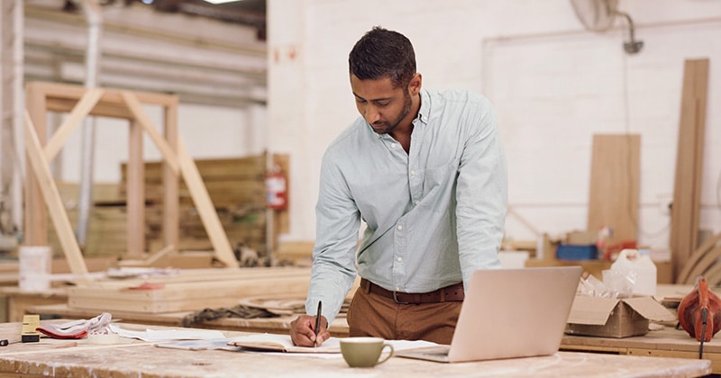 Carpenter in workshop with open laptop and writing in notebook
