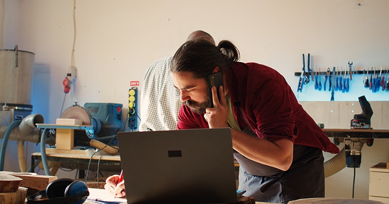 Contractor on phone in front of open laptop in shop