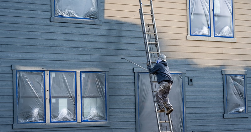 Painting professional in hard hat and air respirator on extension ladder with paint sprayer