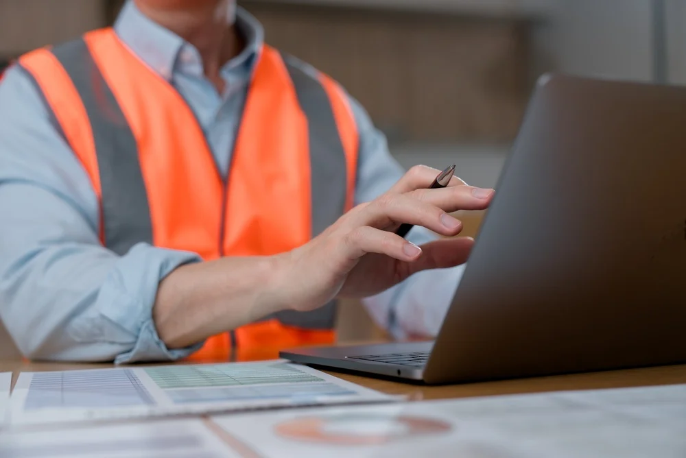 Construction engineer in an orange safety vest using a laptop at a desk with project blueprints.