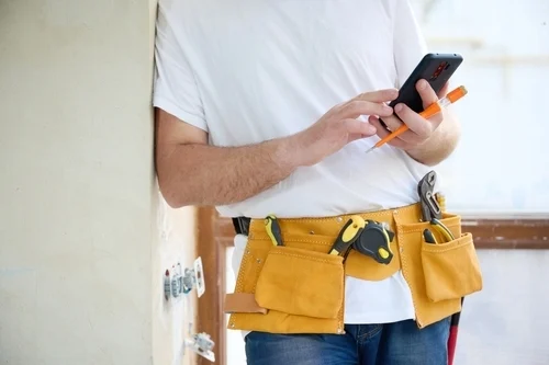 A close-up of a construction worker in a white t-shirt and yellow tool belt using a smartphone.
