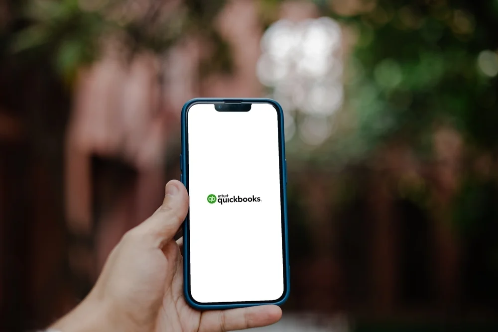 A person holding a smartphone showing the Intuit QuickBooks mobile app splash screen.