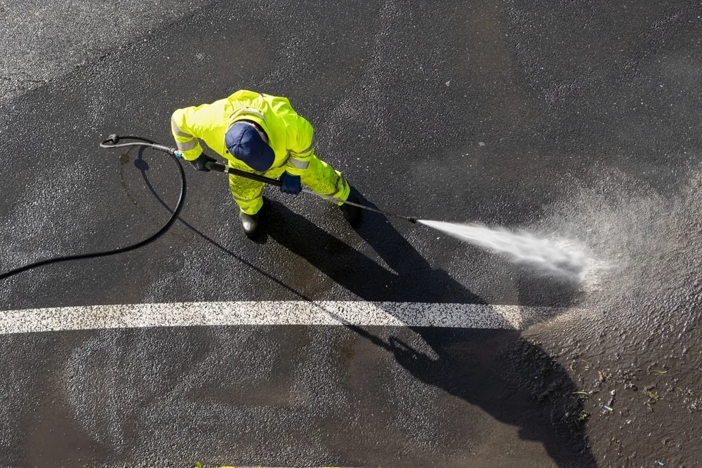 • Worker in high-visibility gear pressure washing an asphalt road.
