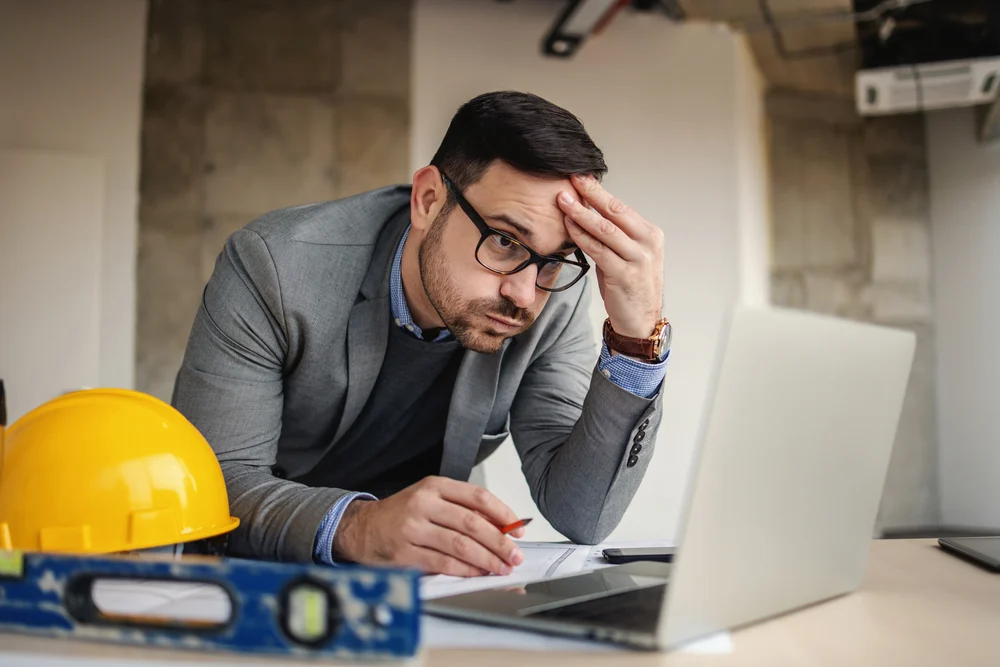 A stressed construction project manager looking at a laptop with blueprints and a yellow hard hat on the desk.