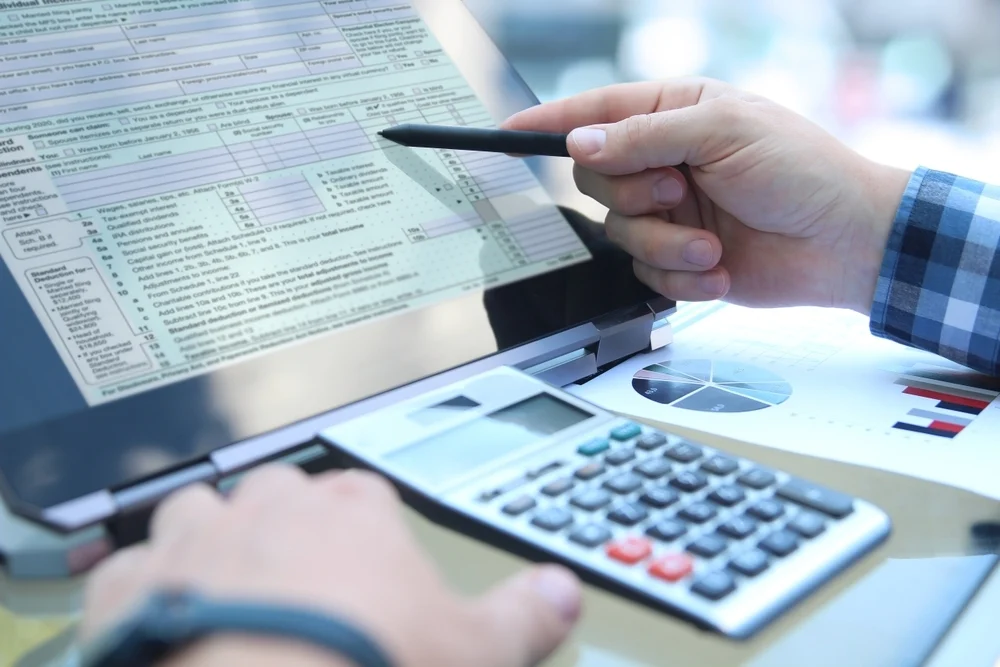 • A person filling out tax return forms on a laptop with a calculator on the desk.
