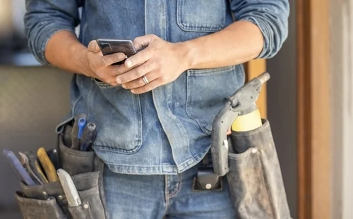 Close-up of a worker in a denim jacket and tool belt using a smartphone.